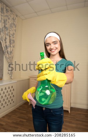 Selected focus on the water sprayer in the hand of beautiful smiling housemaid wearing nice shirt and jeans on background
