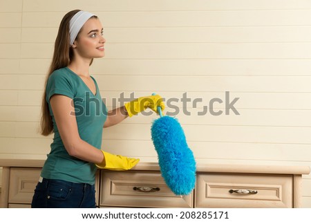 Lovely dark-haired housemaid with the white fillet wearing yellow gloves cleaning cream-colored chest of drawers with the duster