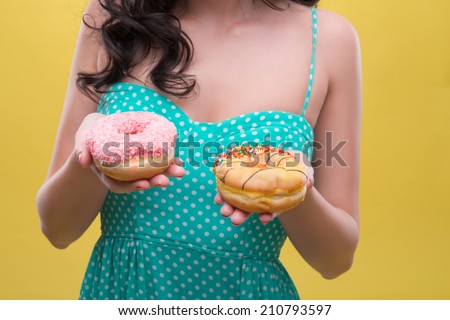 Half-length portrait of dark-haired woman wearing nice mint dotted dress holding two doughnuts and decided which one she wants to eat. Isolated on yellow background
