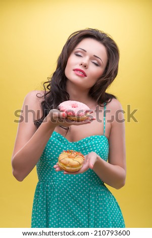 Half-length portrait of dark-haired woman wearing nice mint dotted dress holding two doughnuts and decided which one she wants to eat. Isolated on yellow background