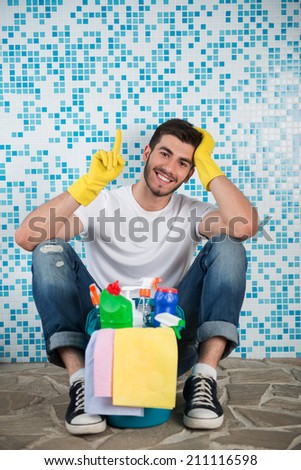 Young smiling dark-haired janitor wearing white shirt blue jeans and yellow rubber gloves sitting in the bathroom with pail of cleaners leaning on the wall satisfied with his work