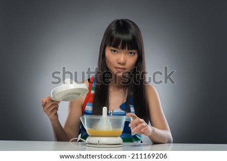 Half-length portrait of very serious young dark-haired Asian woman sitting at the table holding the important part of juicer. Isolated on black background
