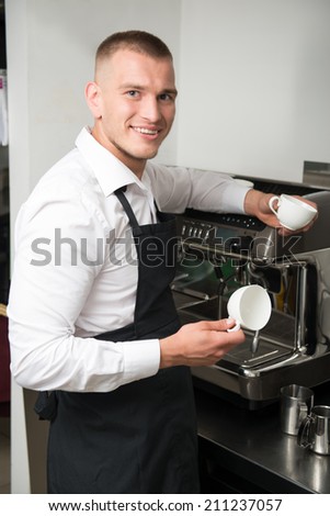 Half-length portrait of handsome young smiling barista wearing white shirt and black apron holding two white cups for coffee standing near coffee machine