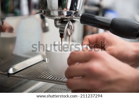 Hands of professional barista holding two white cups on the grating of coffee machine looking how coffee pouring into them