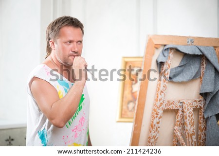 Half-length portrait of fair-haired thoughtful painter wearing white painted shirt standing opposite the picture holding the brush in his mouth and looking at his work