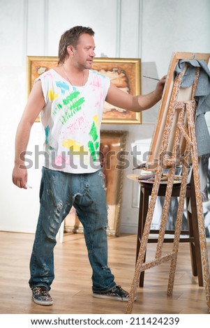 Full-length portrait of handsome fair-haired painter wearing white painted shirt and blue torn jeans drawing his picture in the studio
