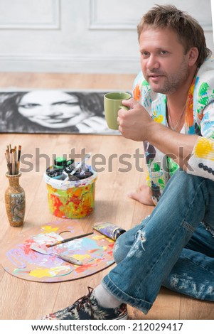 Portrait of thoughtful fair-haired painter wearing cool painted jacket and torn jeans sitting on the floor in his studio near his famous picture having a rest with the cup of tea