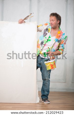 Full-length portrait of fair-haired surprised painter wearing cool painted jacket and torn jeans holding a pot with different paints and brushes looking out of the white linen for copy place
