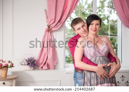 Selected focus on smiling young fair-haired man wearing dark-red shirt and jeans hugging his happy pregnant wife wearing nice sundress. Their living-room on background