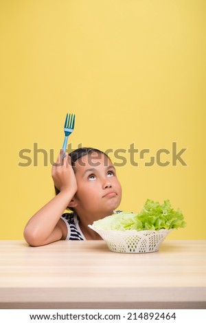 Half-length portrait of pretty dark-haired little girl wearing nice striped dress sitting at the table near the plate of lettuce dreaming about new doll. Isolated on yellow background