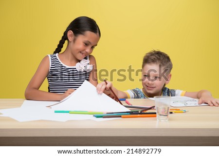 Little smiling fair-haired cute boy sitting at the table with many felt-tips on it and peeping in the drawing of pretty dark-haired girl sitting side by side with him. Isolated on yellow background