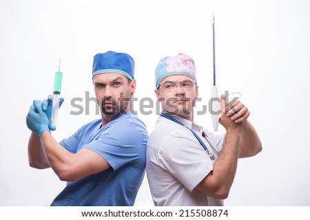 Half-length portrait of two funny doctors wearing medical dress standing back to each other holding injector and medical apparatus like weapon. Isolated on white background