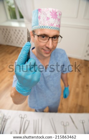 Selective focus on the scalpel in the hands of smiling doctor wearing blue disposable gloves and medical uniform standing on background. Top view