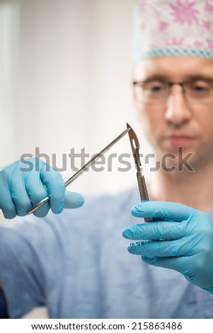 Selective focus on the scalpel and pair of scissors in the hands of the serious doctor wearing blue disposable gloves standing on background