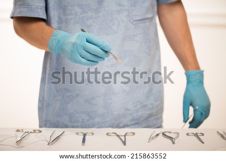 Selective focus on the tweezers in the hands of the surgeon wearing blue disposable gloves standing near the table with surgical tools