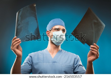 Half-length portrait of the surgeon wearing blue medical uniform and mask holding two radiographs in his hand looking at them very attentively. Isolated on blue background