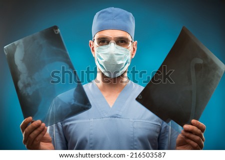Half-length portrait of the surgeon wearing blue medical uniform and mask holding two radiographs in his hands looking at us. Isolated on blue background
