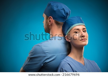 Half-length portrait of two young doctors wearing blue medical uniform standing back to each other with crossing arms isolated on blue background