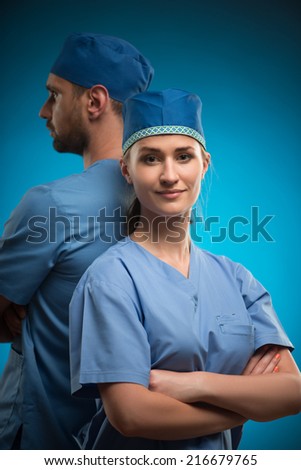 Half-length portrait of two young doctors wearing blue medical uniform standing back to each other with crossing arms isolated on blue background