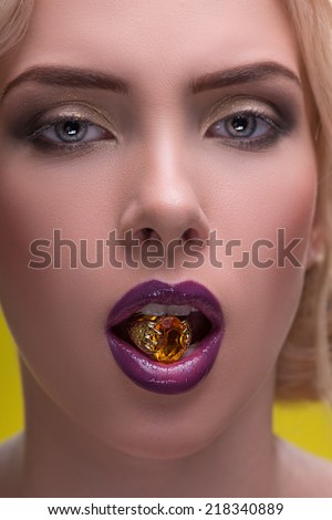 Half-length portrait of beautiful sexy blonde with violet tempting lips and perfect skin holding precious gold fingering in her mouth isolated on yellow background. Close up