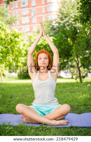 Pretty young red-haired woman wearing white T-shirt and mental shorts doing yoga lotus pose sitting on the blue mat in the park