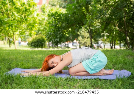 Pretty young red-haired woman wearing white T-shirt and mental shorts doing yoga lying like a ball on the blue mat in the park