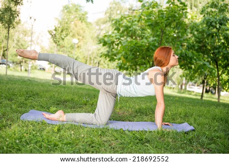 Pretty young red-haired woman wearing white T-shirt and grey pants doing yoga on the blue mat in the park