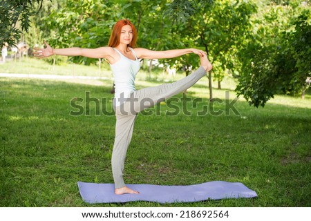 Pretty young red-haired woman wearing white T-shirt and grey pants doing yoga stretching standing on the one hand on blue mat in the park