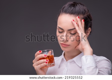 Half-length portrait of dark-haired tired beautiful woman wearing white blouse standing aside with closed eyes holding a glass of whisky thinking about her boyfriend. Isolated on dark background