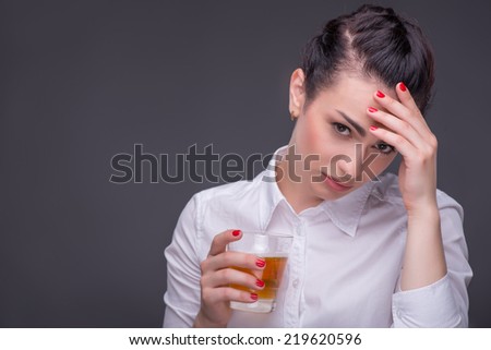 Half-length portrait of dark-haired tired beautiful woman wearing white blouse sitting holding a glass of whisky thinking about her terrible life. Isolated on dark background