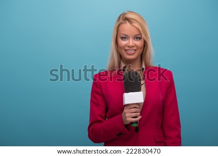 Half-length portrait of very beautiful fair-haired smiling TV presenter wearing great red jacket and cream-colored shirt speaking into the microphone. Isolated on blue background