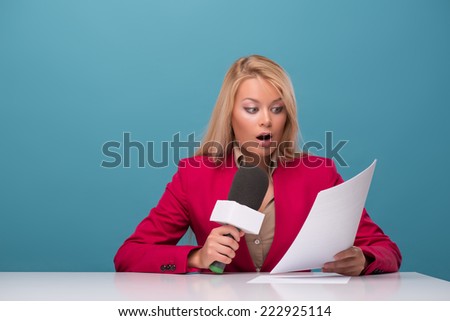 Half-length portrait of good-looking fair-haired surprised TV presenter wearing great red jacket and cream-colored shirt sitting at the table reading fresh news. Isolated on blue background