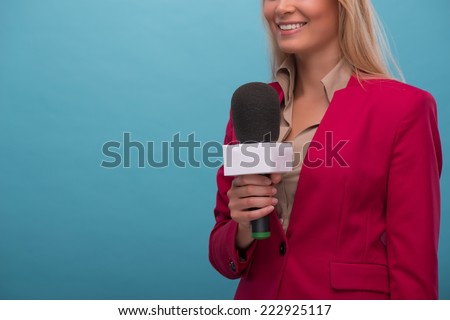 Half-length portrait of very beautiful fair-haired TV presenter wearing great red jacket and cream-colored shirt standing aside with the microphone. Isolated on blue background