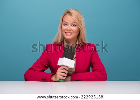 Half-length portrait of good-looking fair-haired smiling TV presenter wearing great red jacket and cream-colored shirt sitting at the table holding a microphone. Isolated on blue background