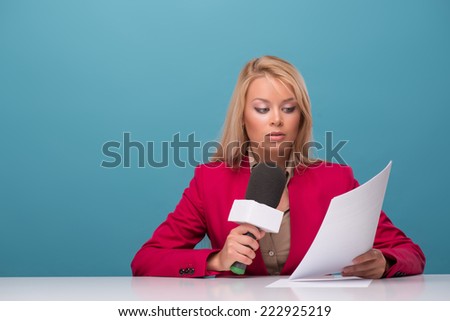Half-length portrait of good-looking fair-haired surprised TV presenter wearing great red jacket and cream-colored shirt sitting at the table reading fresh news. Isolated on blue background