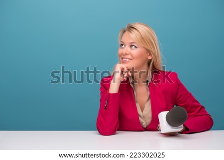 Half-length portrait of lovely fair-haired TV presenter wearing great red jacket and cream-colored shirt sitting at the table holding a microphone dreaming about something wonderful. Isolated on blue