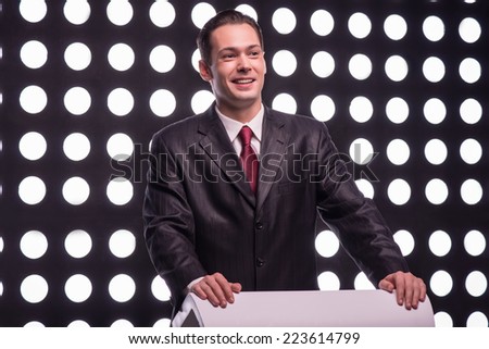 Half- length portrait of young handsome smiling TV presenter wearing great black suit and vinous tie standing behind the rostrum announcing the opening speech