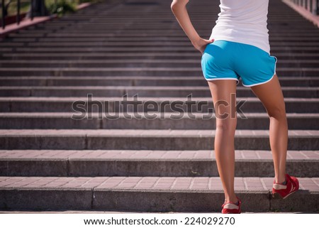 Selective focus on the young sexy woman wearing white T-shirt and blue shorts standing back in the red jogging shoes on the stairs on background