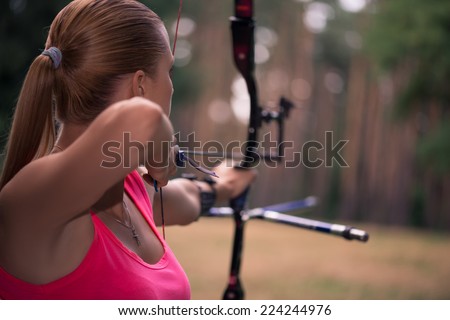 Selective focus on the lovely young fair-haired woman standing back wearing pink T-shirt pulling the bowstring