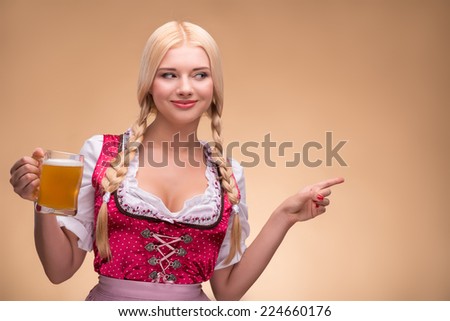 Young sexy smiling blonde wearing pink dirndl with white blouse holding a beer mag looking and pointing at someone. Isolated on dark background