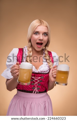 Half-length portrait of young sexy surprised blonde wearing pink dirndl and white blouse holding in both hands beer mugs looking at us. Isolated on dark background