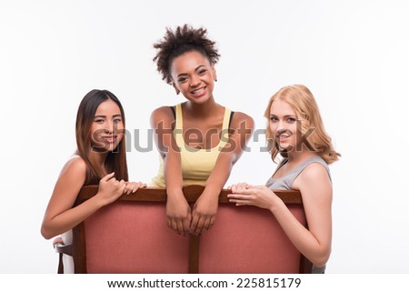 Half-length portrait of three pretty girls wearing jeans and T-shirts sitting on the sofa having fun and laughing. Isolated on white background