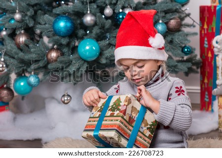 Half-length portrait of the little cute fair-haired girl sitting on the floor near Christmas presents wearing warm sweater and red cap of Santa Claus wanted to unpack her nice present