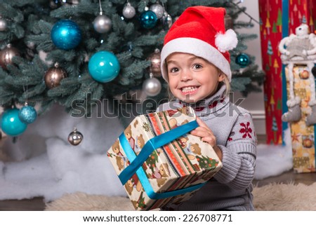 Half-length portrait of the little cute fair-haired smiling girl sitting on the floor near Christmas presents wearing warm sweater and red cap of Santa Claus wanted to unpack her nice present