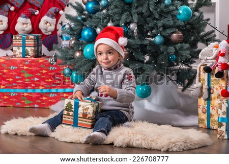Half-length portrait of the little cute fair-haired smiling girl sitting on the floor near Christmas presents wearing warm sweater and red cap of Santa Claus unpacking her nice present