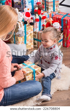 Half-length portrait of beautiful smiling mom and daughter wearing warm sweaters and jeans sitting aside near the Christmas tree holding nice present talking to each other