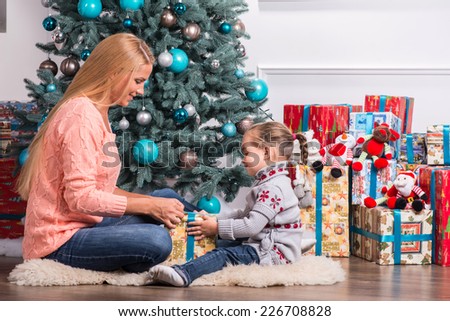 Half-length portrait of beautiful smiling mom and daughter wearing warm sweaters and jeans sitting aside near the Christmas tree holding nice present talking to each other