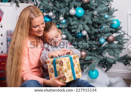Half-length portrait of lovely mom and little daughter wearing warm sweaters sitting near the Christmas tree with wonderful present from Santa Claus