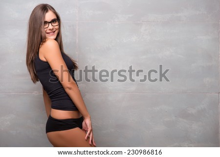 Half-length portrait of sexy smiling dark -haired young woman in nice eyeglasses wearing black vest and shorts standing aside showing her perfect figure