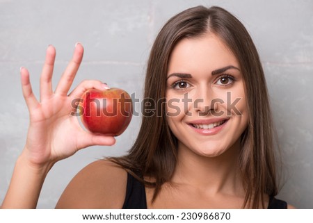 Half-length portrait of sexy smiling dark -haired young woman wearing black vest holding very delicious ripe apple suggested us to taste it